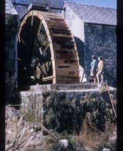 Exterior view of pair of stone watermill buildings with single very large wheel between them
